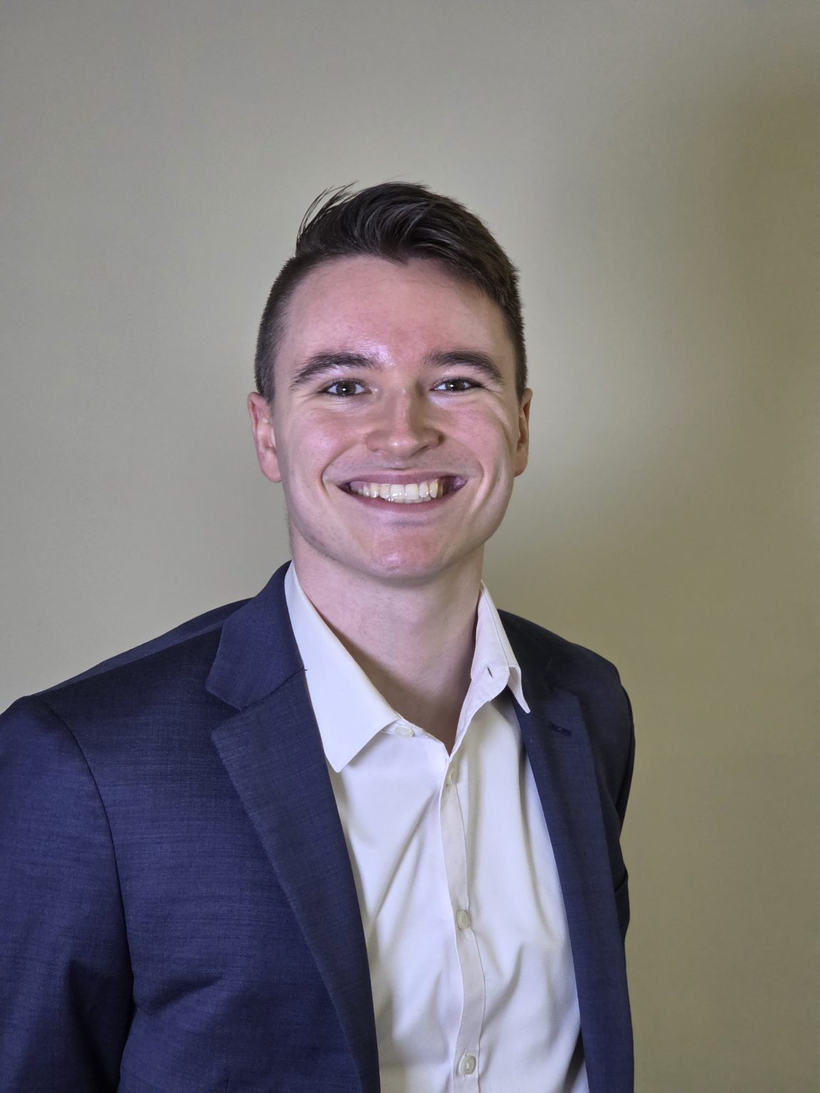Professional headshot of the founder of Pregi Systems, a smiling young man with short dark hair, wearing a navy blue blazer over a white button-down shirt, against a plain, light beige background.
